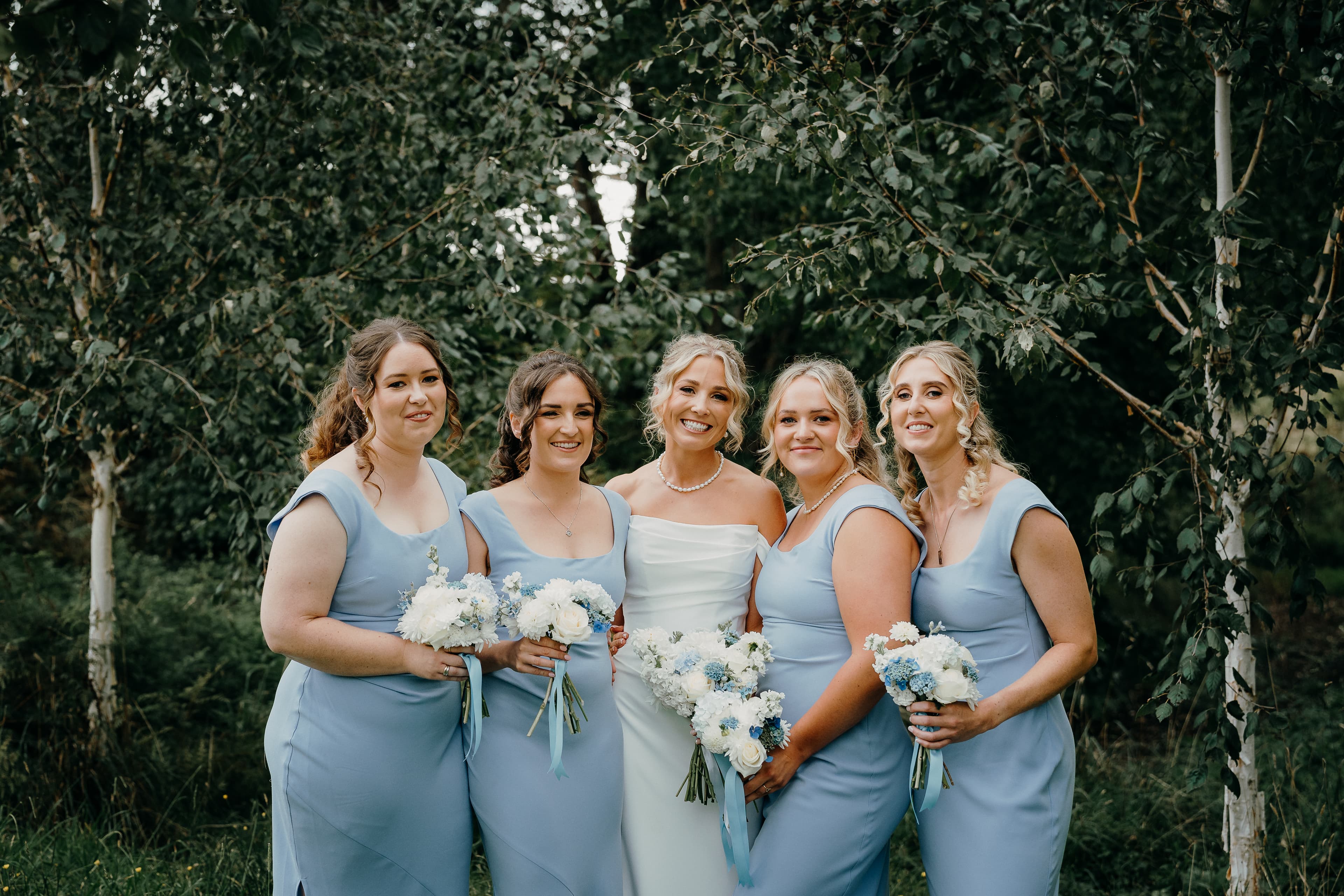 Bride and four bridesmaids in dusty blue dresses smiling together in front of trees, holding white and blue bouquets
